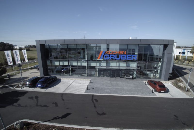 Moderner Glasbau mit „Möbel Gruber“-Schild, Parkplatz mit einigen Autos und wehende Fahnen unter klarem Himmel.