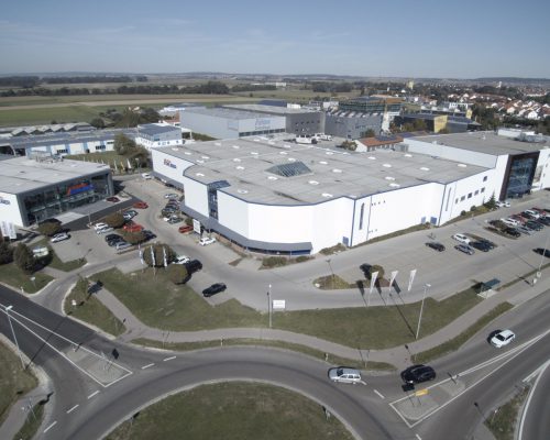 Luftaufnahme des Industriekomplexes von Möbel Gruber mit weißen Gebäuden, umgeben von Parkplätzen und Straßen in der Nähe einer Kleinstadt.