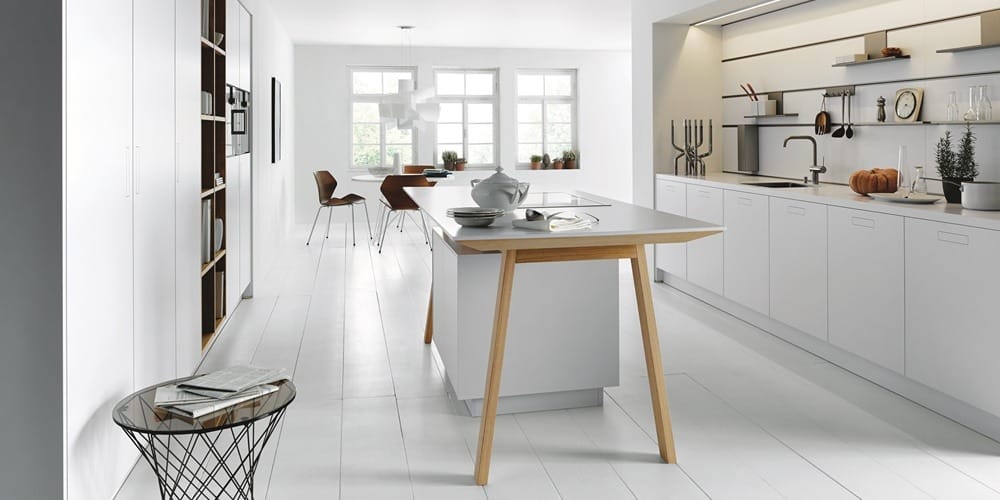Moderne Küche mit weißen Schränken von Möbel Gruber, einer Holzinsel und einem Beistelltisch mit Drahtgestell. Helles Licht fällt durch große Fenster.