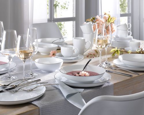 Elegante Tischdekoration von Möbel Gruber mit weißem Geschirr, Weingläsern und Suppenschüsseln auf einer hellgrauen Tischdecke in der Nähe großer Fenster.
