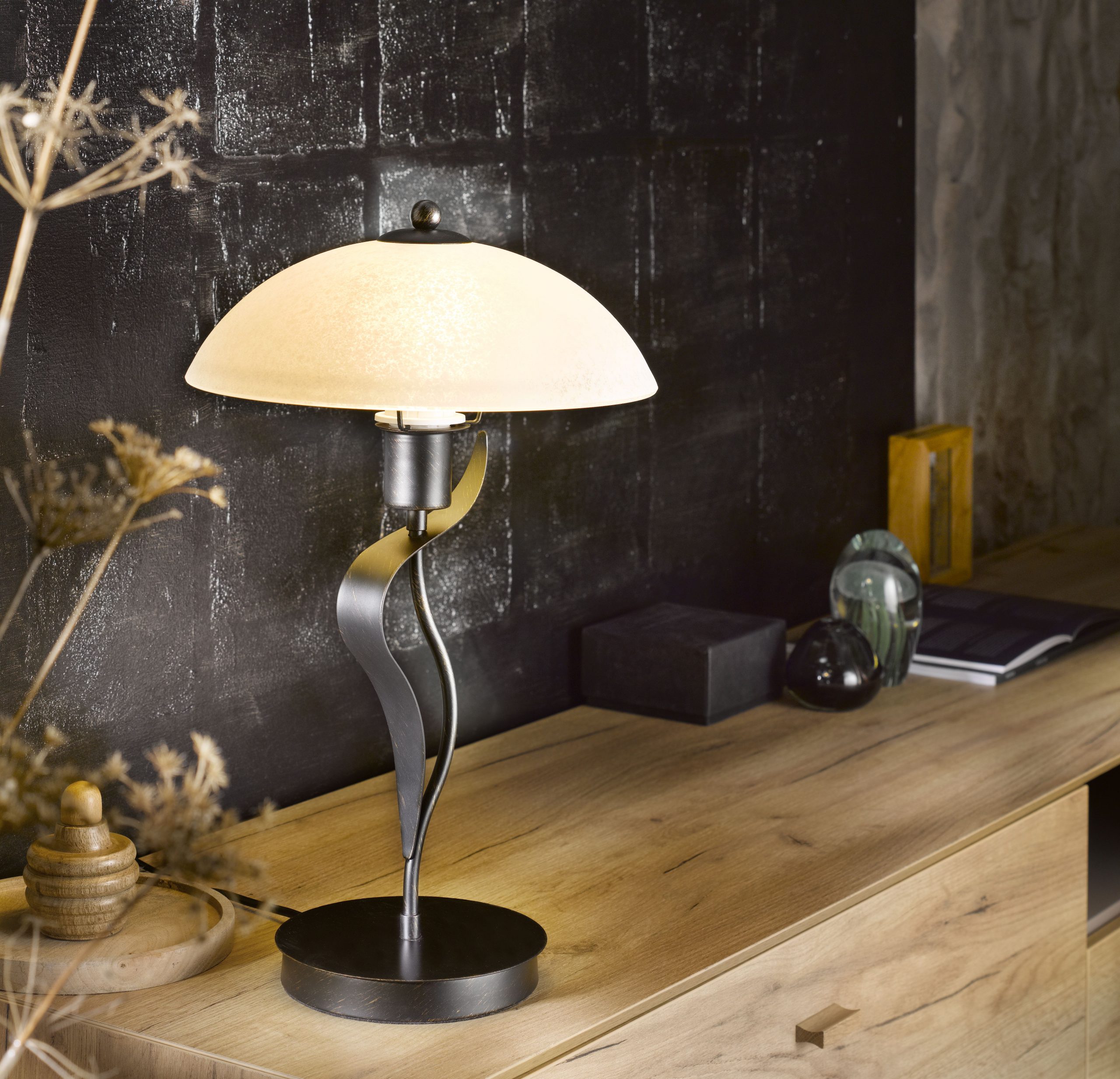 Eine moderne Tischlampe von Möbel Gruber mit geschwungenem Metallsockel und weißem Schirm ziert ein Holzsideboard vor einer strukturierten schwarzen Wand.