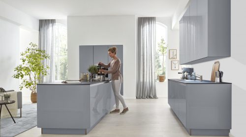 Eine Person bereitet Kaffee in einer modernen Küche von Möbel Gruber mit grauen Schränken, großen Fenstern und Grünpflanzen zu.