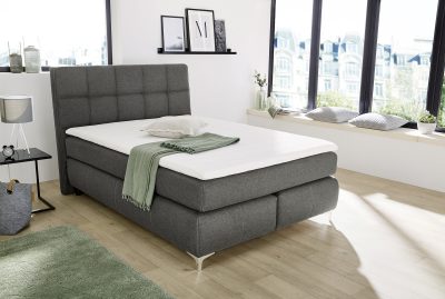 Ein modernes Schlafzimmer von Möbel Gruber präsentiert das Boxspringbett mit weißer Bettwäsche, einer grünen Überwurfdecke und Blick auf die Stadt.