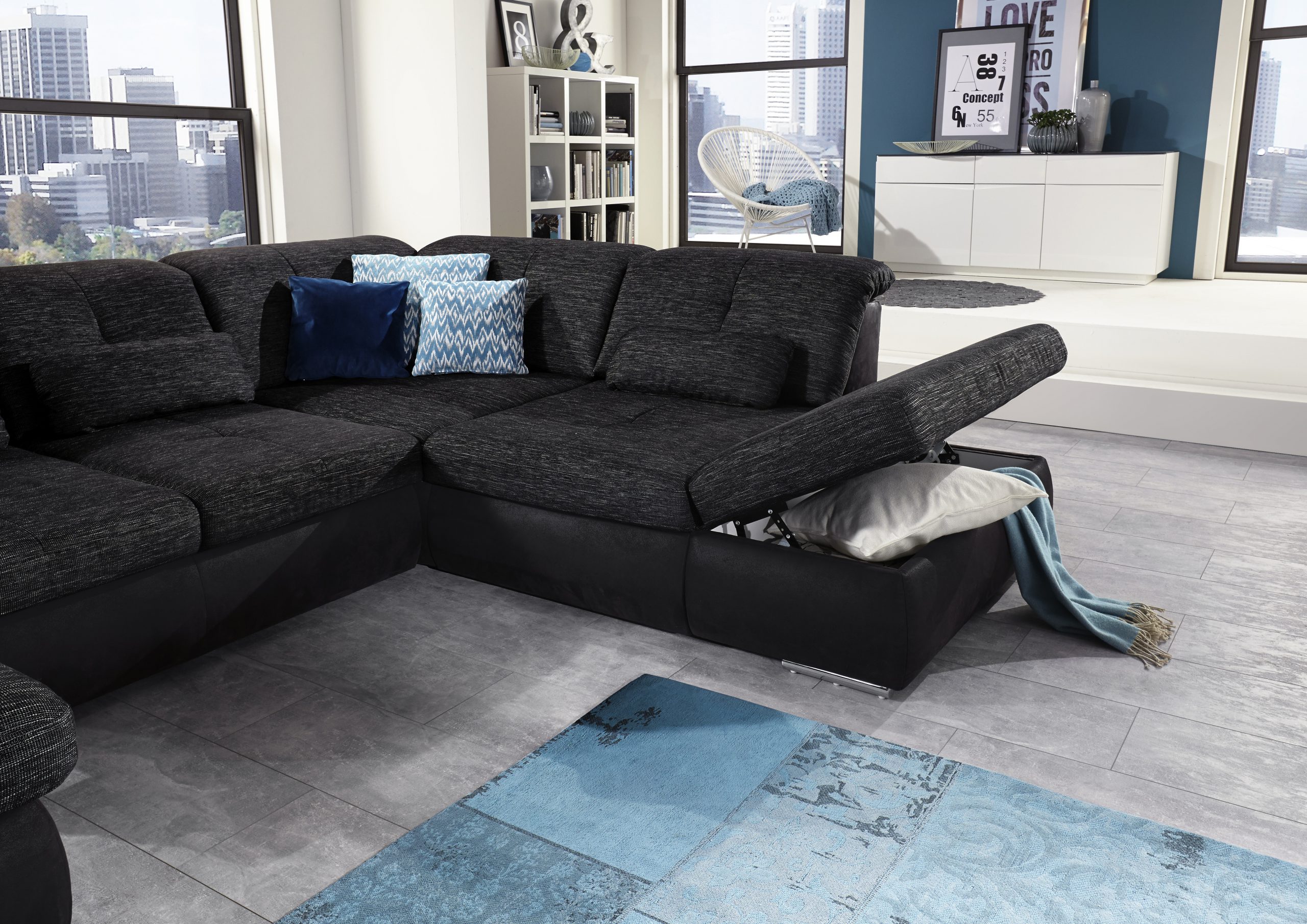 Ein modernes Wohnzimmer von Möbel Gruber mit einem dunkelgrauen Sofa, Kissen, einer blauen Überwurfdecke, weißen Regalen und Blick auf die Stadt.