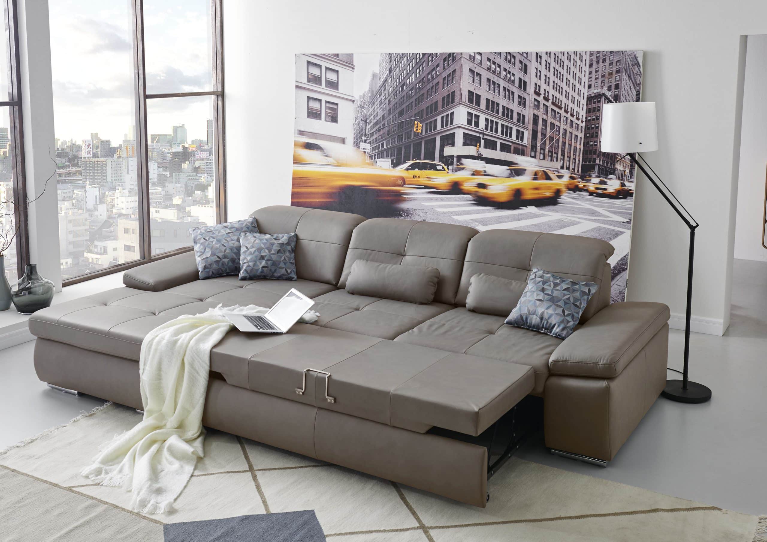 Ein modernes Wohnzimmer von Möbel Gruber mit einem grauen Sofa, einer gemütlichen Überwurfdecke, Kissen, einem Laptop und einem Kunstwerk mit Stadtansichten.