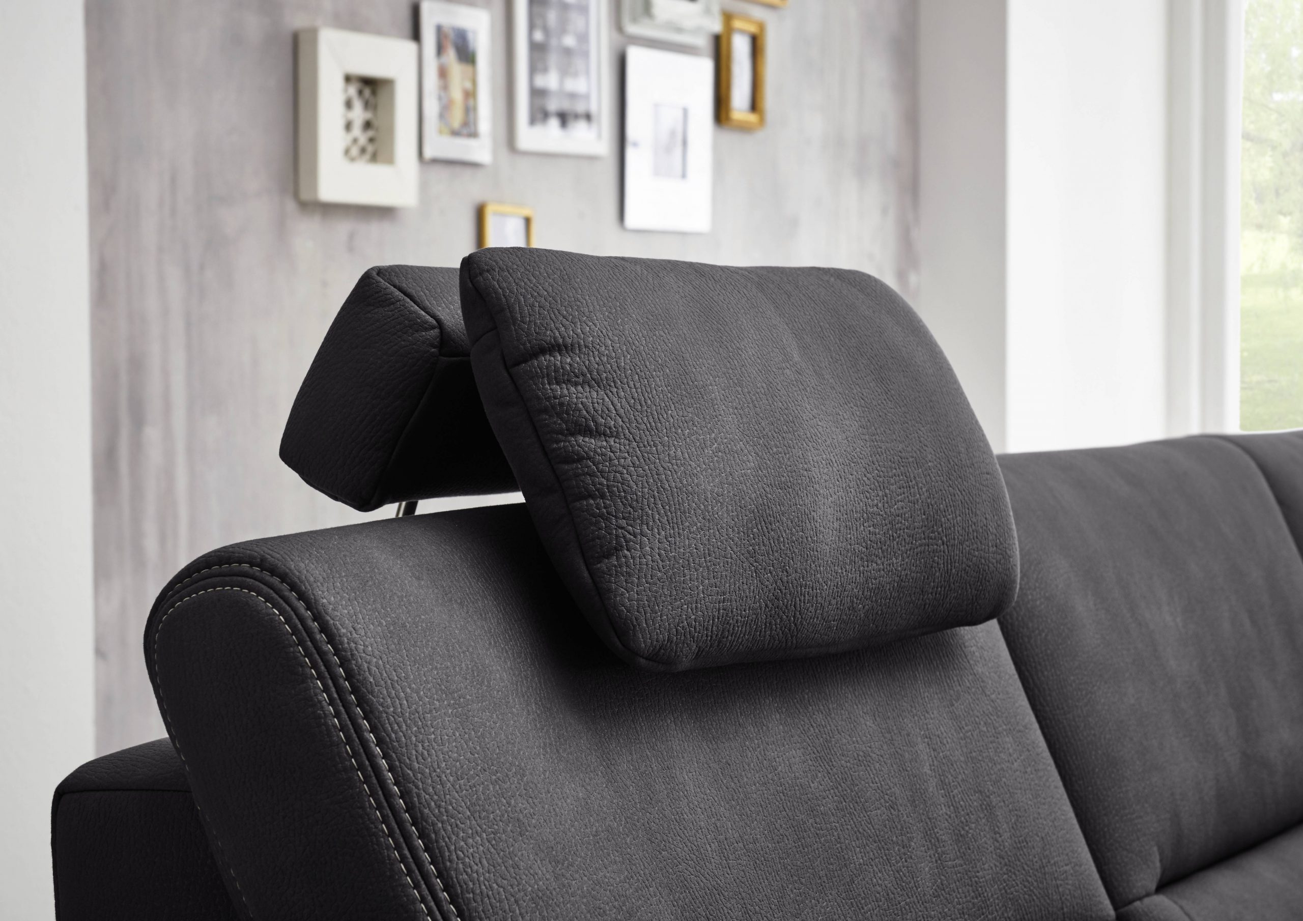 Nahaufnahme eines schwarzen Ledersofas von Möbel Gruber mit verstellbarer Kopfstütze. Unscharfe gerahmte Bilder schmücken die hellgraue Wand.