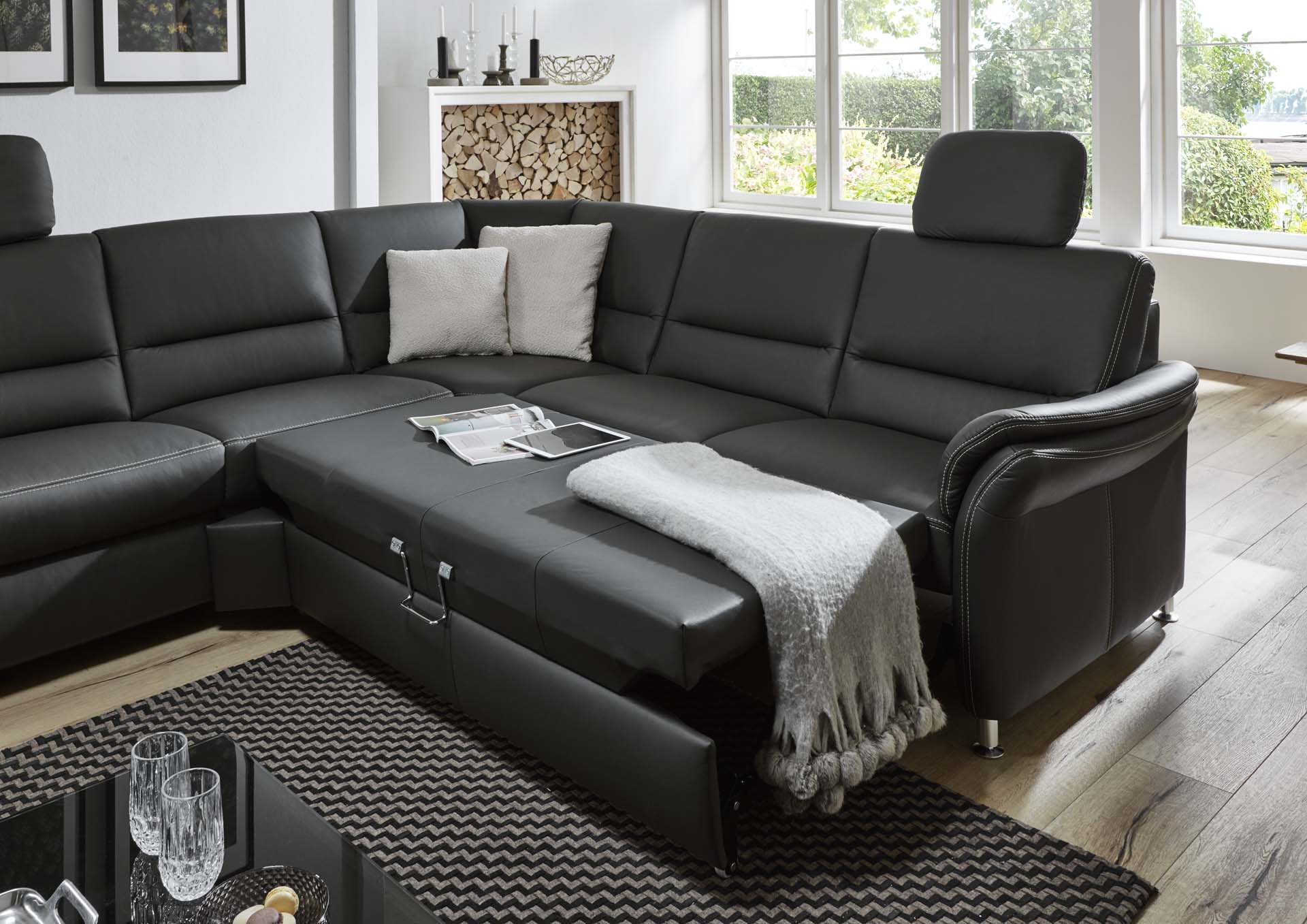 Modernes Wohnzimmer mit schwarzem Sofa von Möbel Gruber, Kuscheldecke, Kissen und einem ordentlich gestapelten Holzstapel am Fenster.