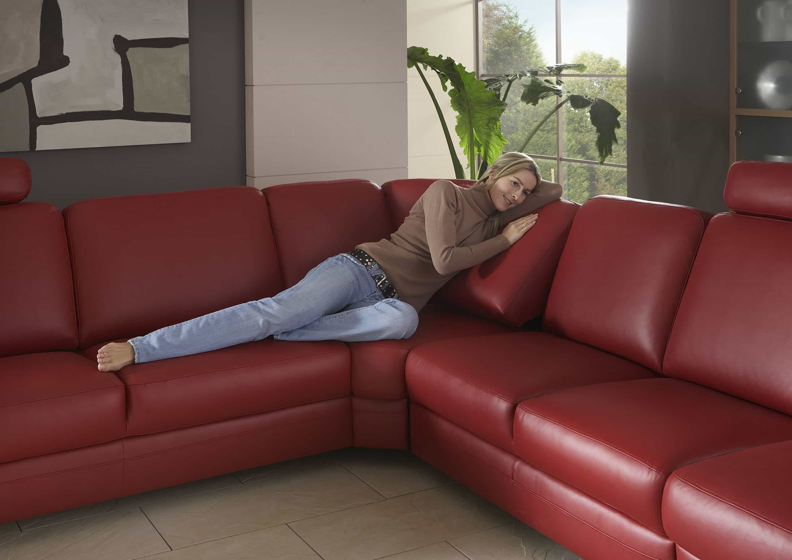 Frau entspannt auf einem roten Sofa von Möbel Gruber in einem modernen Wohnzimmer mit großen Fenstern und einer Blattpflanze.
