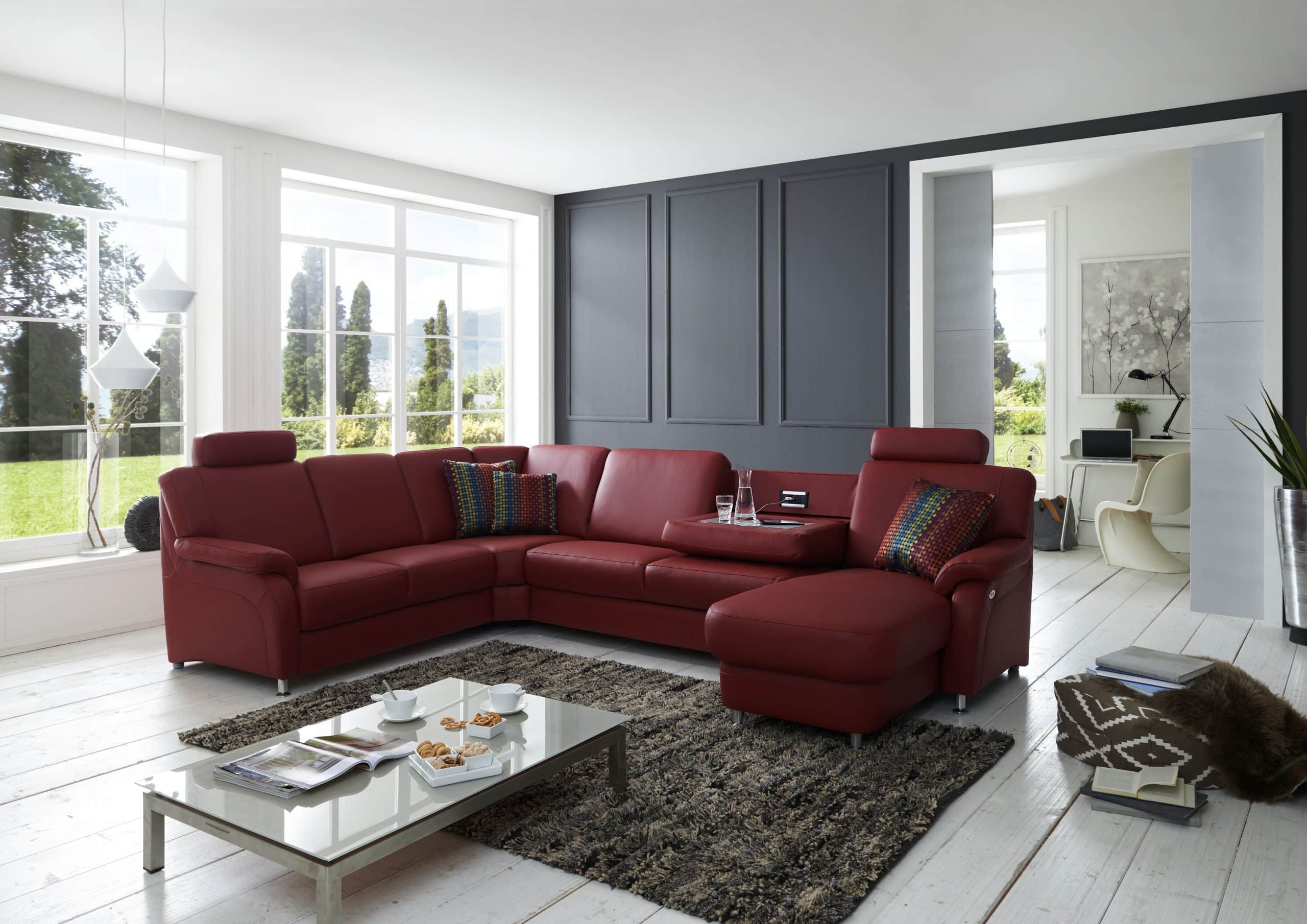 Modernes Wohnzimmer von Möbel Gruber mit rotem Sofa, grauer Akzentwand und großen Fenstern mit viel Grün.