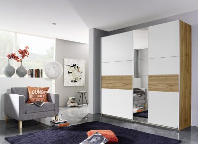 Ein modernes Wohnzimmer von Möbel Gruber mit Schwebetürenschrank, einem grauen Sofa, dekorativen Kissen, Kunst und einem Hochflorteppich.