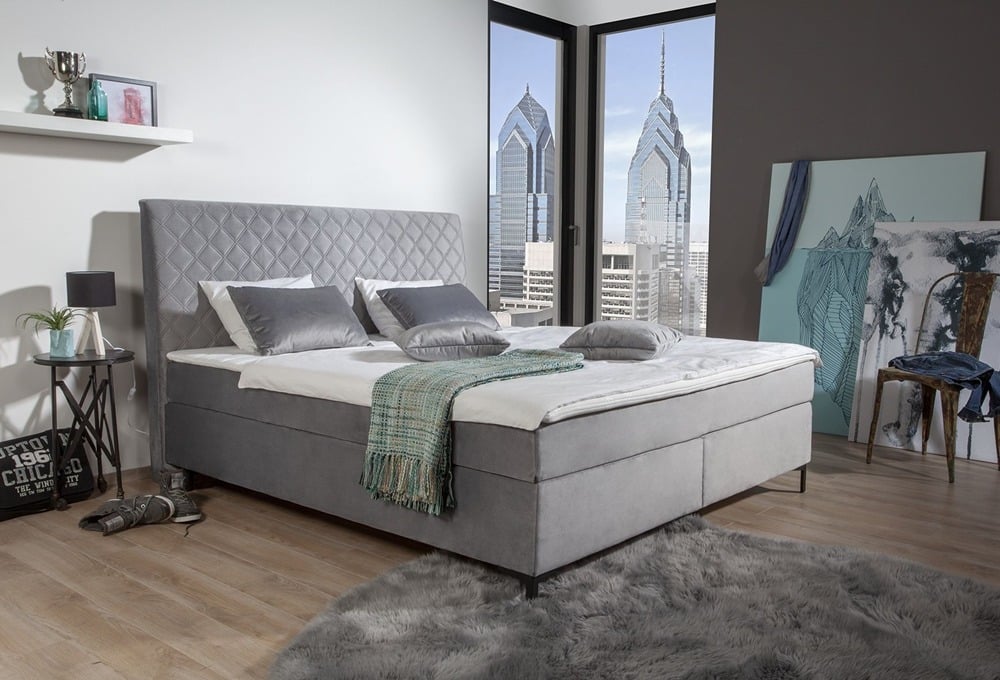 Modernes Schlafzimmer mit grau gepolstertem Bett von Möbel Gruber, Kissen, blaugrüner Überdecke, Blick auf die Wolkenkratzer der Stadt und gemütlichem grauen Teppich.