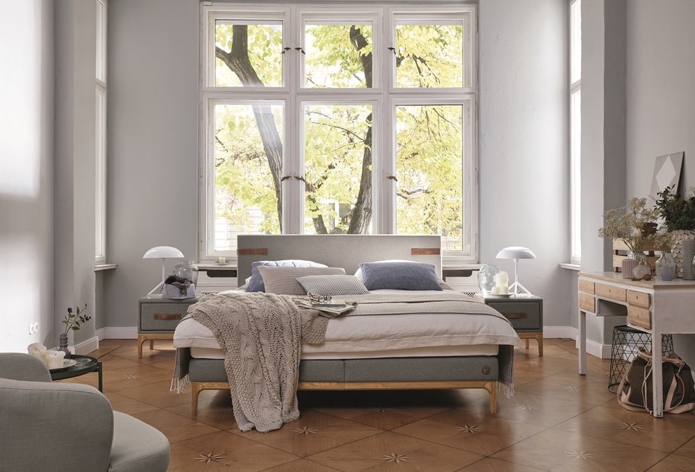 Gemütliches Schlafzimmer von Möbel Gruber mit großen Fenstern, einem gepolsterten Bett, Nachttischen, einem Stuhl und einem blumengeschmückten Schreibtisch.