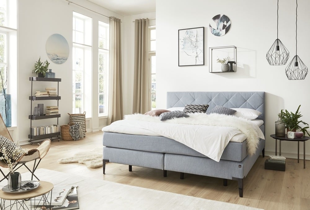 Modernes Schlafzimmer von Möbel Gruber mit hellblauem Bett, weißer Bettwäsche und dekorativer Beleuchtung. Mit Pflanzen, Bücherregalen und Wandkunst.