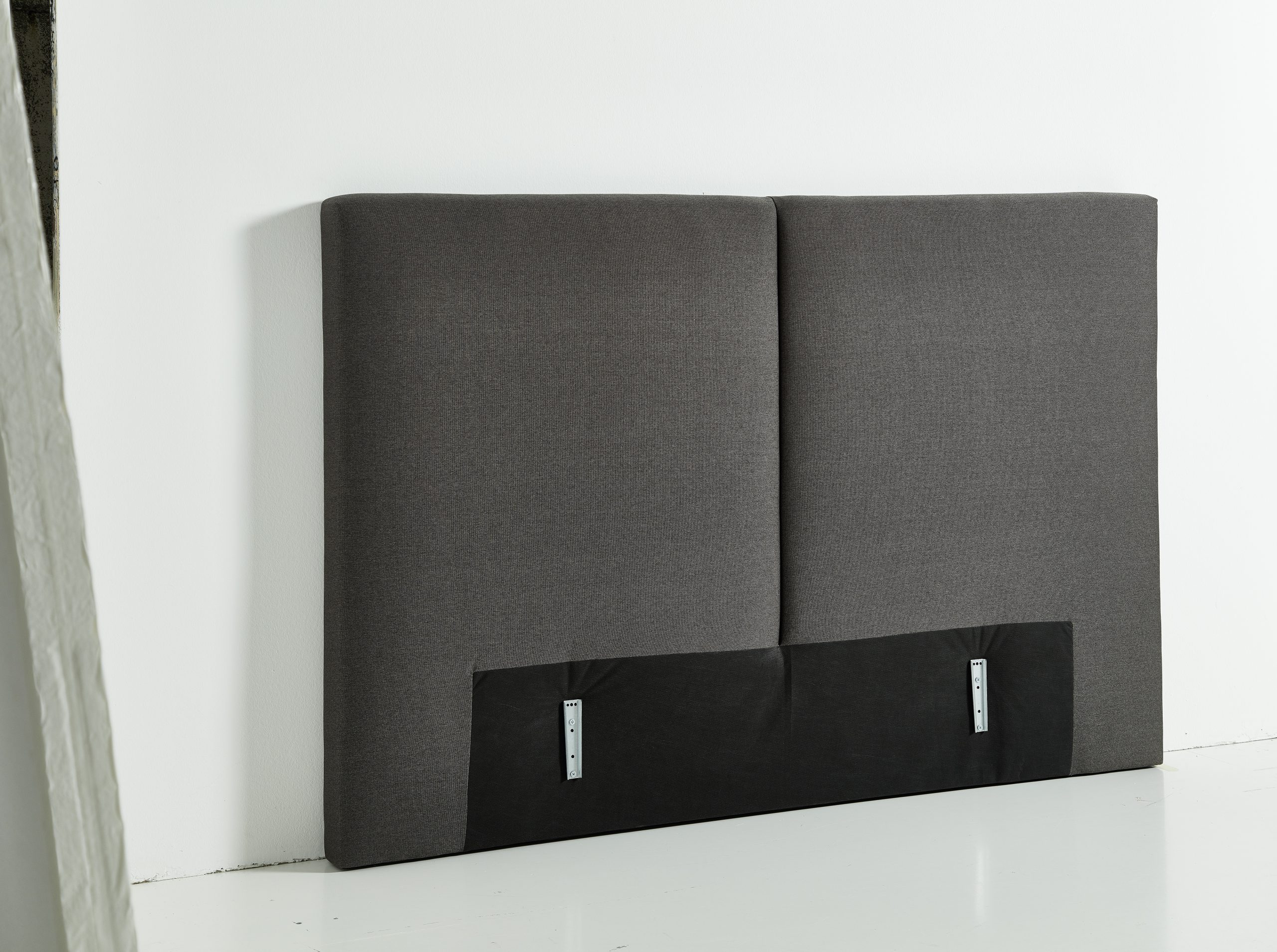 Das grau gepolsterte Kopfteil mit schwarzer Blende von Möbel Gruber ruht elegant vor einer weißen Wand.