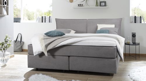 Modernes Schlafzimmer mit einem grau gepolsterten Bett von Möbel Gruber, weißer Bettwäsche, grünen Akzenten und minimalistischem Dekor.