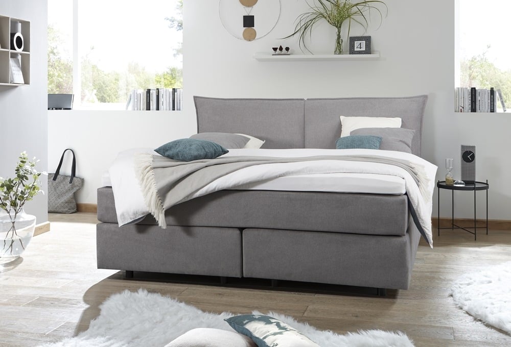 Modernes Schlafzimmer mit einem grau gepolsterten Bett von Möbel Gruber, weißer Bettwäsche, blaugrünen Akzenten und minimalistischem Dekor.
