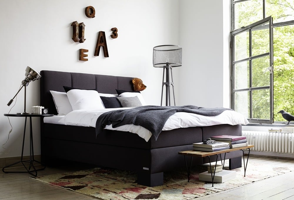 Modernes Schlafzimmer von Möbel Gruber mit dunklem Polsterbett, dekorativen Wandbuchstaben, großem Fenster und minimalistischer Einrichtung.