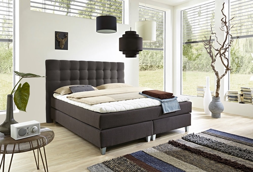 Modernes Schlafzimmer von Möbel Gruber mit großem Bett, grauem Kopfteil, abstraktem Dekor, großen Fenstern, üppigen Pflanzen und eleganter Beleuchtung.