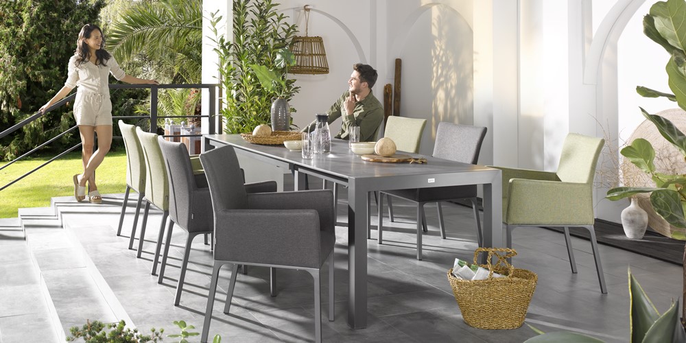 Ein Mann und eine Frau genießen ein Gespräch auf einer Terrasse mit modernem Esstischset von Möbel Gruber, umgeben von üppigem Grün.