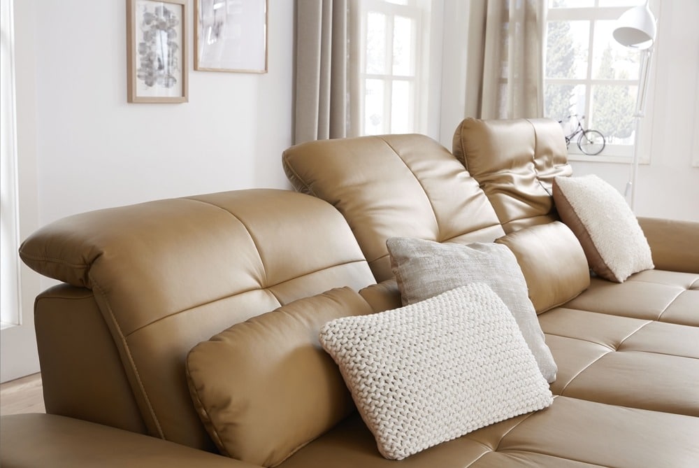 Hellbraunes Ledersofa von Möbel Gruber mit neutralen Kissen in einem hellen Wohnzimmer mit gerahmter Kunst und großen Fenstern.
