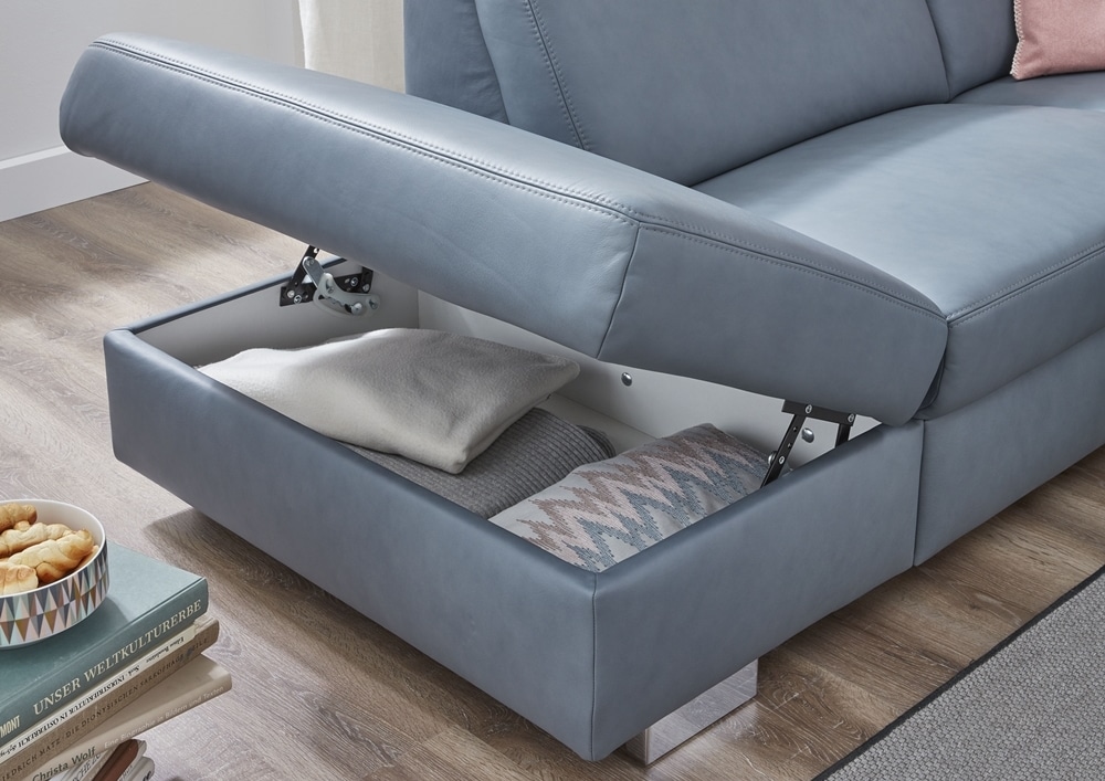 Entdecken Sie den versteckten Stauraum des Sofas von Möbel Gruber, der mit ordentlich gefalteten Decken gefüllt ist. In der Nähe bietet ein Couchtisch Snacks an.