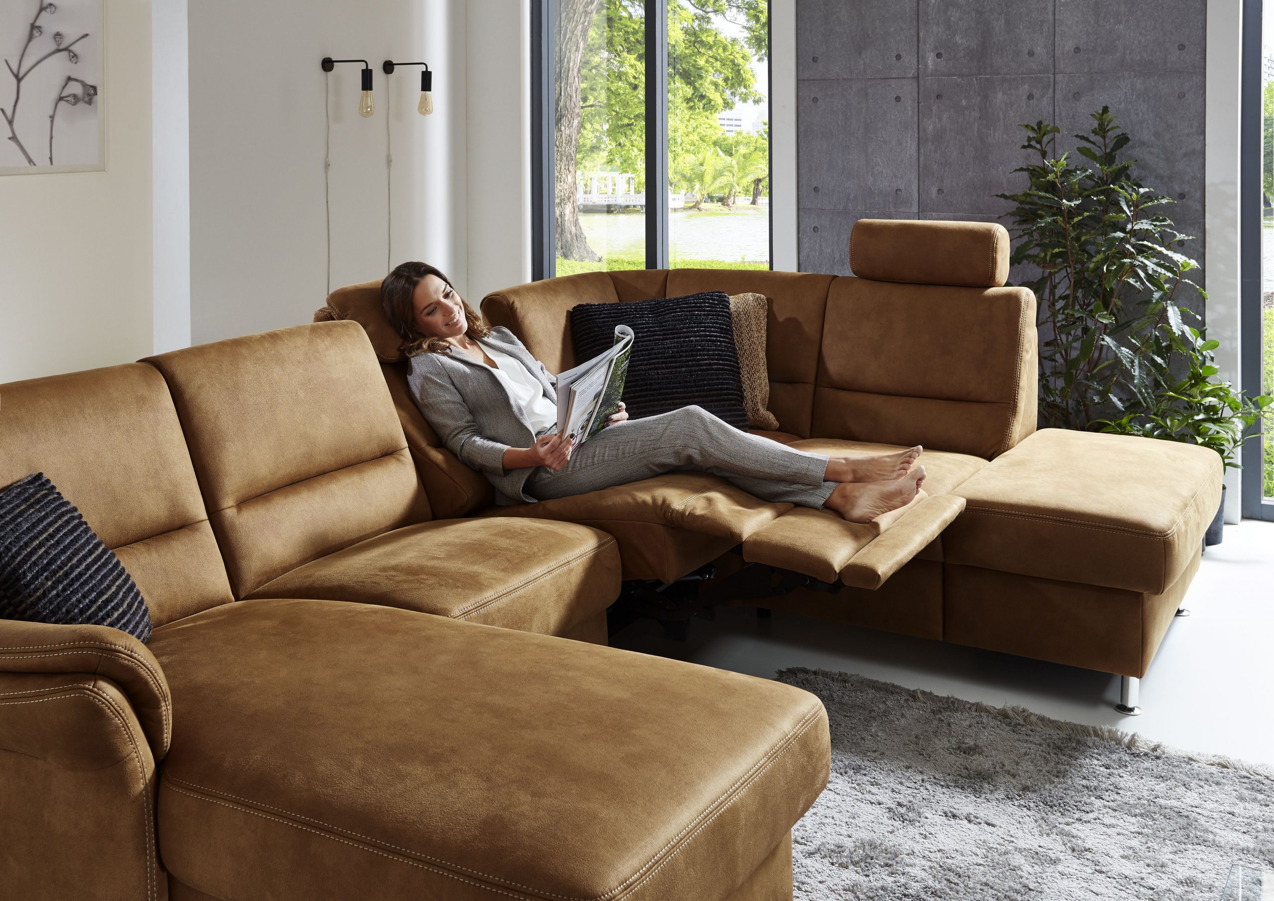 Eine Person entspannt sich auf einem Sofa von Möbel Gruber und liest eine Zeitschrift in einem hellen Raum mit großen Fenstern und einer Pflanze in der Nähe.