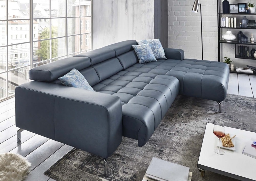 Modernes Wohnzimmer mit einem dunkelblauen Sofa von Möbel Gruber, gemusterten Kissen, einer Stehlampe und einem Couchtisch mit einem Weinglas.