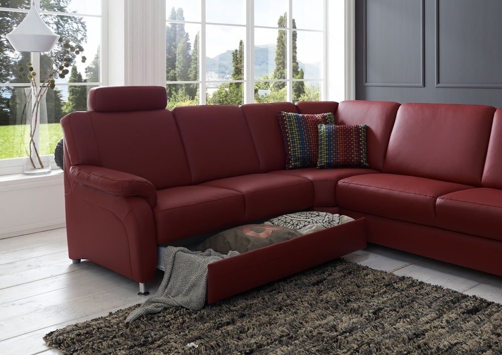 Rotes Ecksofa von Möbel Gruber mit Kissen, einer ausziehbaren Schublade auf einem grauen Teppich. Große Fenster rahmen den Blick auf Bäume ein.