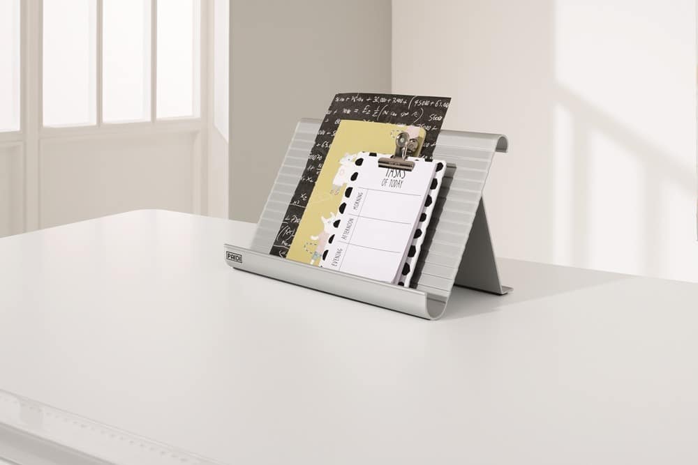 Ein eleganter Metall-Organizer von Möbel Gruber auf einem weißen Schreibtisch hält Papiere und einen Planer vor einer hellen, durch ein Fenster beleuchteten Wand.