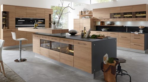 Moderne Küche von Möbel Gruber mit Holzschränken, Kochinsel, Barhocker und Pflanzen, alles in natürliches Licht getaucht.