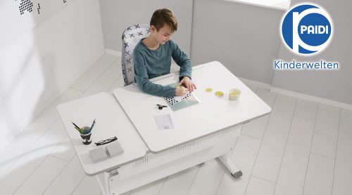 Kind sitzt an einem weißen Verstellschreibtisch von Möbel Gruber und macht Hausaufgaben. Daneben stehen Schreibwaren und eine Tasse. In der Ecke das Logo „Paidi Kinderwelten“.
