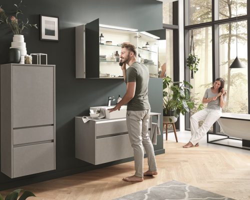 Ein Mann an einem Waschbecken von Möbel Gruber telefoniert, während eine Frau in der Badewanne eines hellen, modernen Badezimmers entspannt.