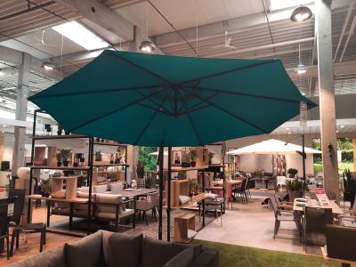 Der Ampelschirm in groß Petrol ist mit verschiedenen Sitzgelegenheiten im Showroom von Möbel Gruber ausgestellt.