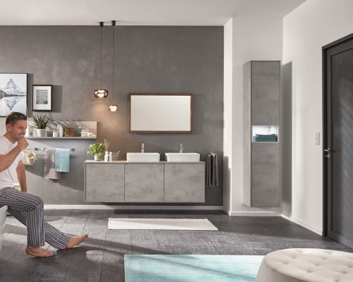 Mann sitzt auf der Badewanne in einem modernen grauen Badezimmer und hält eine Kaffeetasse; im Hintergrund Waschbecken, Spiegel und Wandkunst von Möbel Gruber.