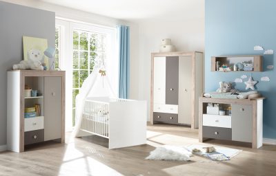 Das helle Babyzimmer von Möbel Gruber verfügt über ein Kinderbett, Holzmöbel, Spielzeug, eine blaue Akzentwand und ein großes Fenster für natürliches Licht.
