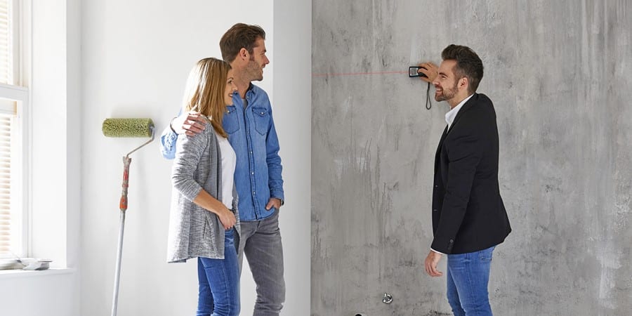 Ein Paar steht mit einem lächelnden Mann vor einer kahlen Wand und hält eine Laserwasserwaage an. An einer anderen Wand lehnt eine Farbrolle.