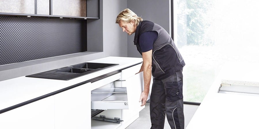 Ein Mann installiert eine Schublade in einer modernen Küche mit weißen Schränken und einer schwarzen Küchenrückwand.