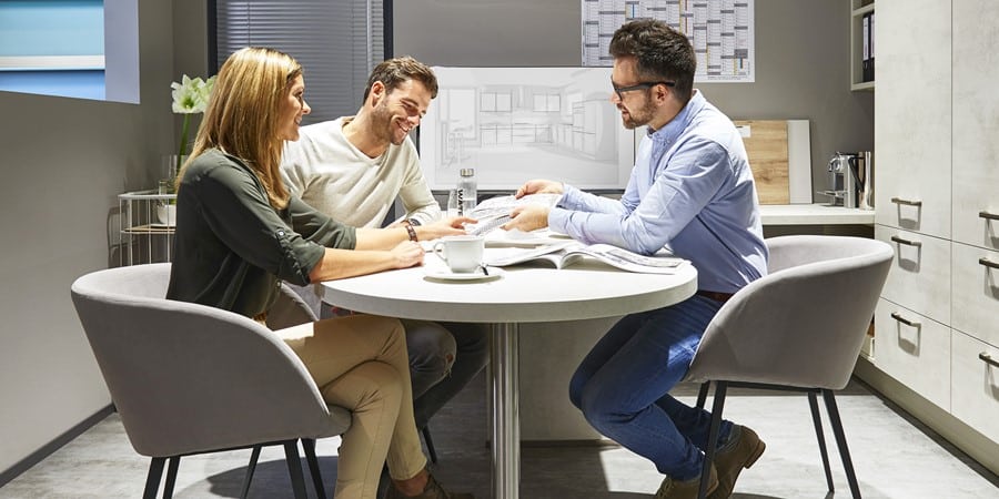 Drei Personen sitzen um einen Tisch und besprechen Pläne in einem Büro, im Hintergrund sind Entwurfsskizzen zu sehen.