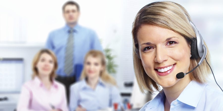 Im Fokus steht eine lächelnde Frau mit Headset, im Hintergrund sind drei verschwommene Personen in Business-Kleidung zu sehen.