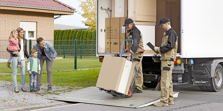 Möbelpacker laden Kisten aus einem Lastwagen aus, während eine Familie mit zwei Kindern in der Nähe zusieht.