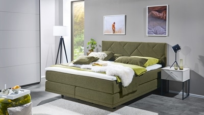 Modernes Schlafzimmer mit einem grünen Polsterbett, grüner Bettwäsche und Kissen. Weißer Nachttisch mit Lampe daneben.