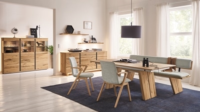 Ein modernes Esszimmer mit Holzmöbeln, blauem Teppich und großen Fenstern. Natürliches Licht durchflutet Tisch, Stühle und Bank.