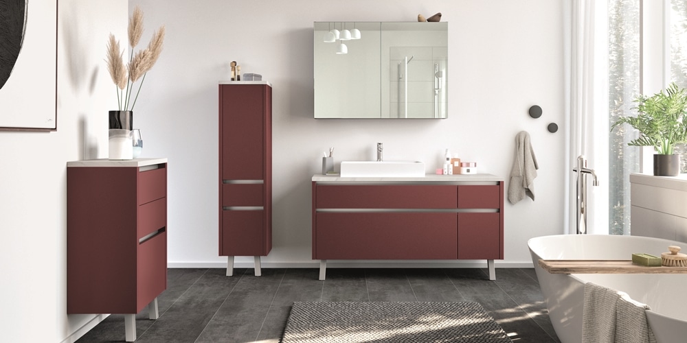 Modernes Badezimmer von Möbel Gruber mit roten Schränken, großem Spiegel, Waschbecken, Badewanne, dekorativen Pflanzen, heller Beleuchtung und neutralen Tönen.