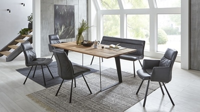 Modernes Esszimmer mit Holztisch von Möbel Gruber, fünf grauen Stühlen, passender Bank und großem Fenster. An der Wand hängt ein Gemälde.