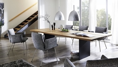 Modernes Esszimmer von Möbel Gruber mit einem Holztisch, sechs grauen Stühlen und zwei Deckenleuchten; dahinter Treppe und Fenster.