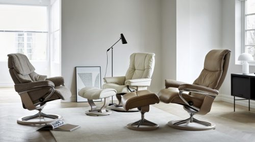 Ein modernes Wohnzimmer von Möbel Gruber mit drei Ledersesseln, einem passenden Fußhocker, einer Stehlampe und sanftem Licht aus einem Fenster.