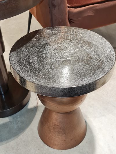 Ein kleiner, runder, metallischer Beistelltisch mit strukturierter Oberfläche auf einem glatten Betonboden.