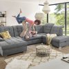 Ein Paar spielt fröhlich auf einem grauen Sofa von Möbel Gruber in einem hellen Wohnzimmer mit großen Fenstern und modernem Dekor.