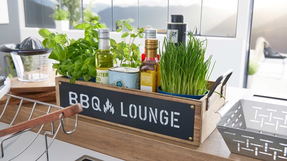 Eine Holzbox mit der Aufschrift „BBQ Lounge“ von Möbel Gruber beherbergt Kräuter, Öle und Gewürze auf der Arbeitsplatte.