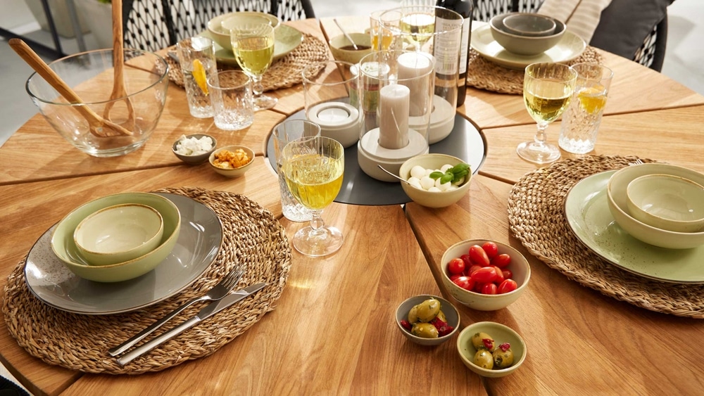 Ein runder Holztisch von Möbel Gruber, gedeckt für eine Mahlzeit mit Geschirr, Gläsern, Tomaten, Oliven und Gewürzen auf gewebten Tischsets.